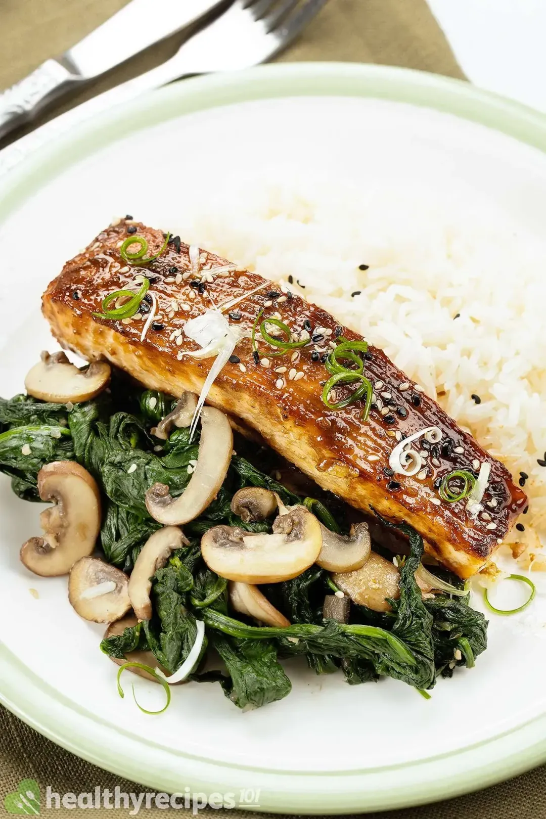 A plate containing a piece of salmon glazed with miso sauce, served alongside cremini mushroom slices, cooked spinach, and white rice