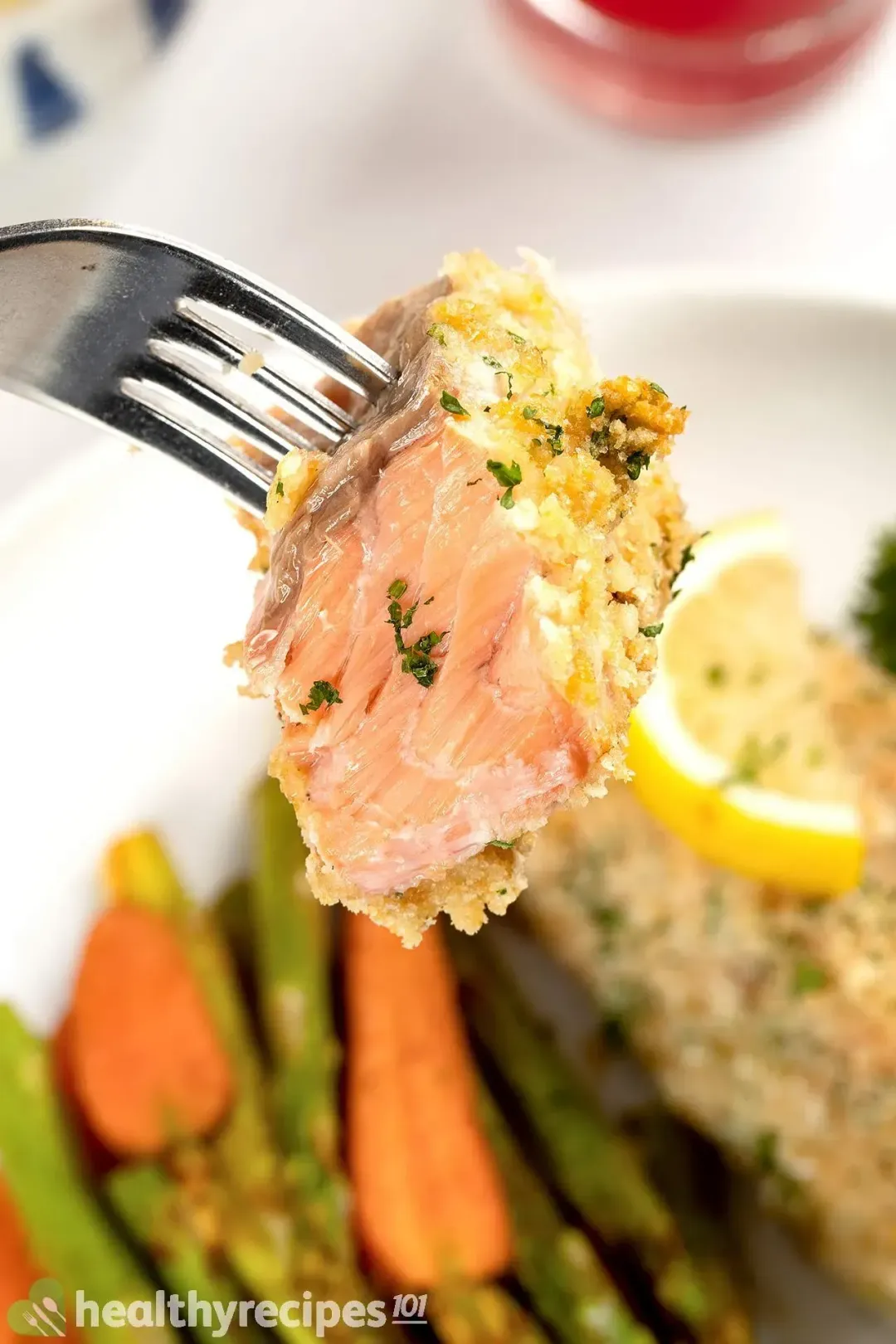 A fork lifting a pale orange piece of breadcrumb-crusted salmon with carrots and asparagus in the blurred background