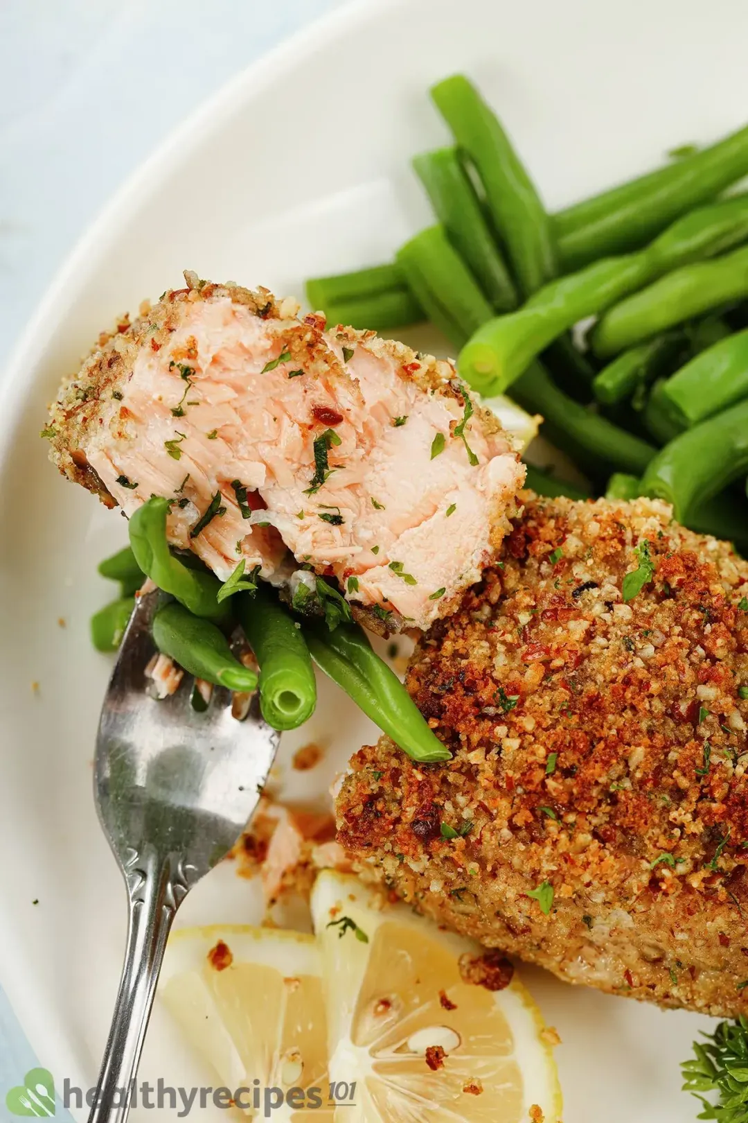 One mouth-sized piece of pecan-crusted salmon is cut from the whole filet and pierced through by a fork; on the same plate placed cooked green beans and lemon slices.