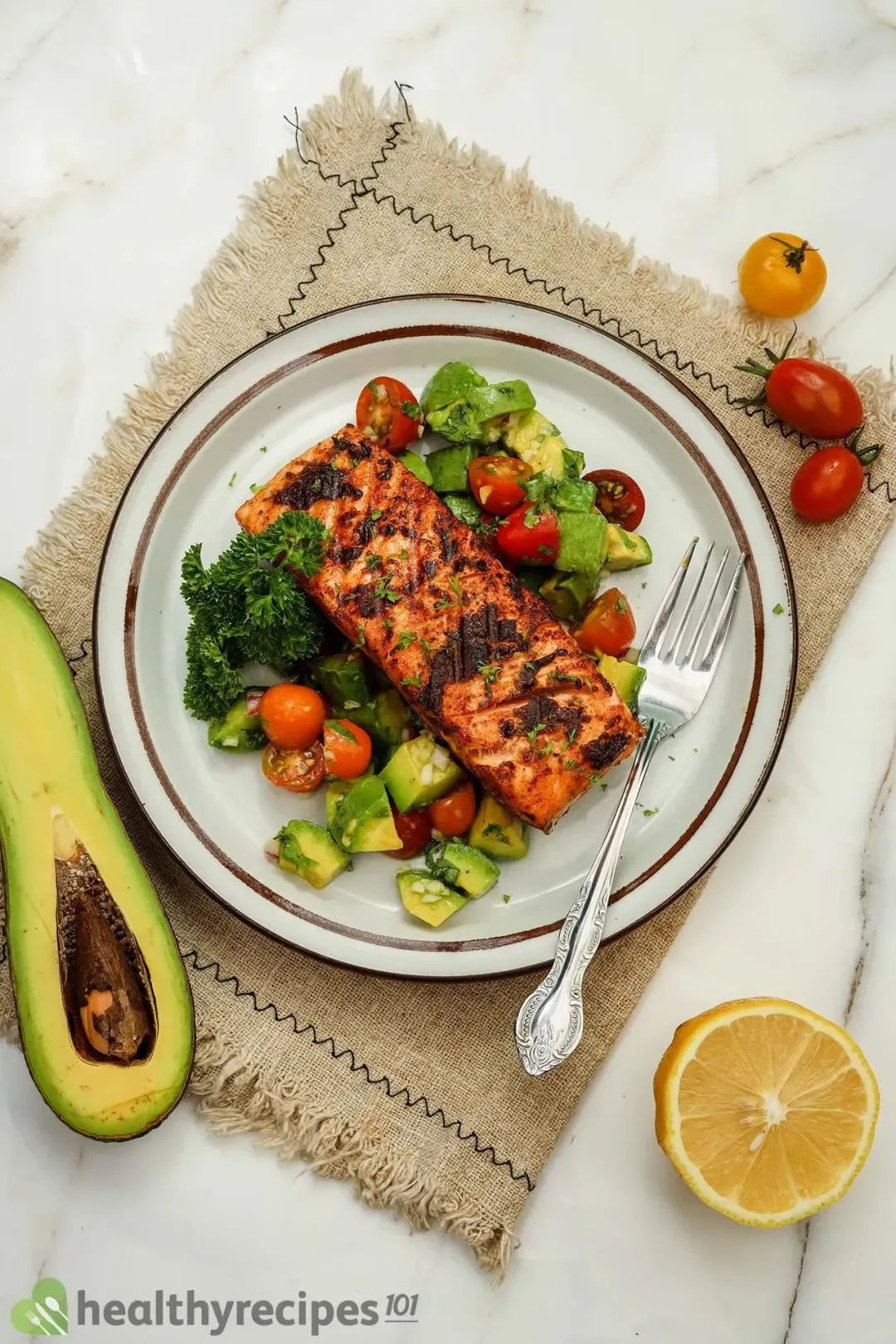A plate of salmon avocado decorated with a fork on a tablecloth surrounded by avocado, lemon, and cherry tomatoes