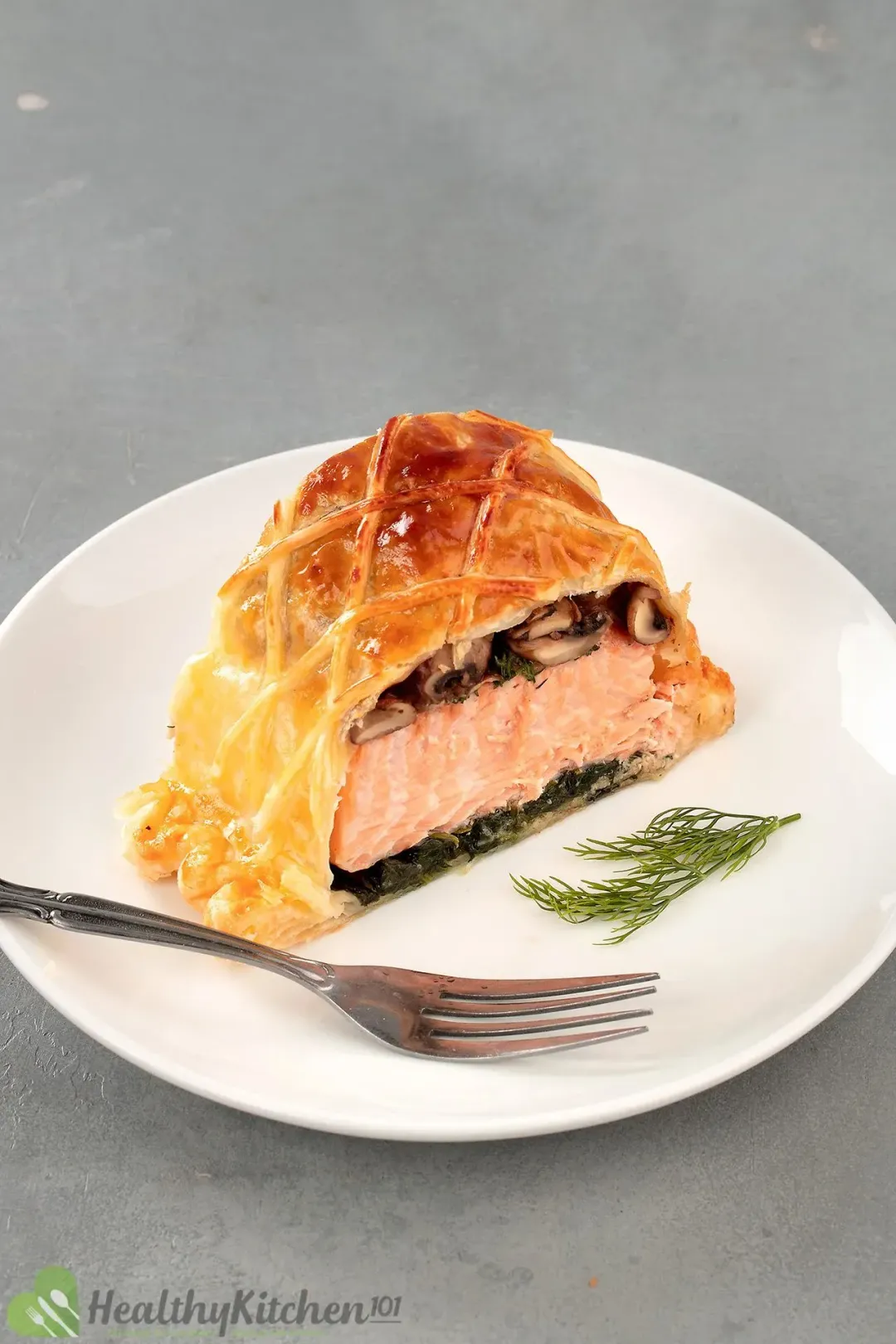 A slice of salmon en croute filled with pink salmon and cooked mushroom laid next to a fork and a fresh dill