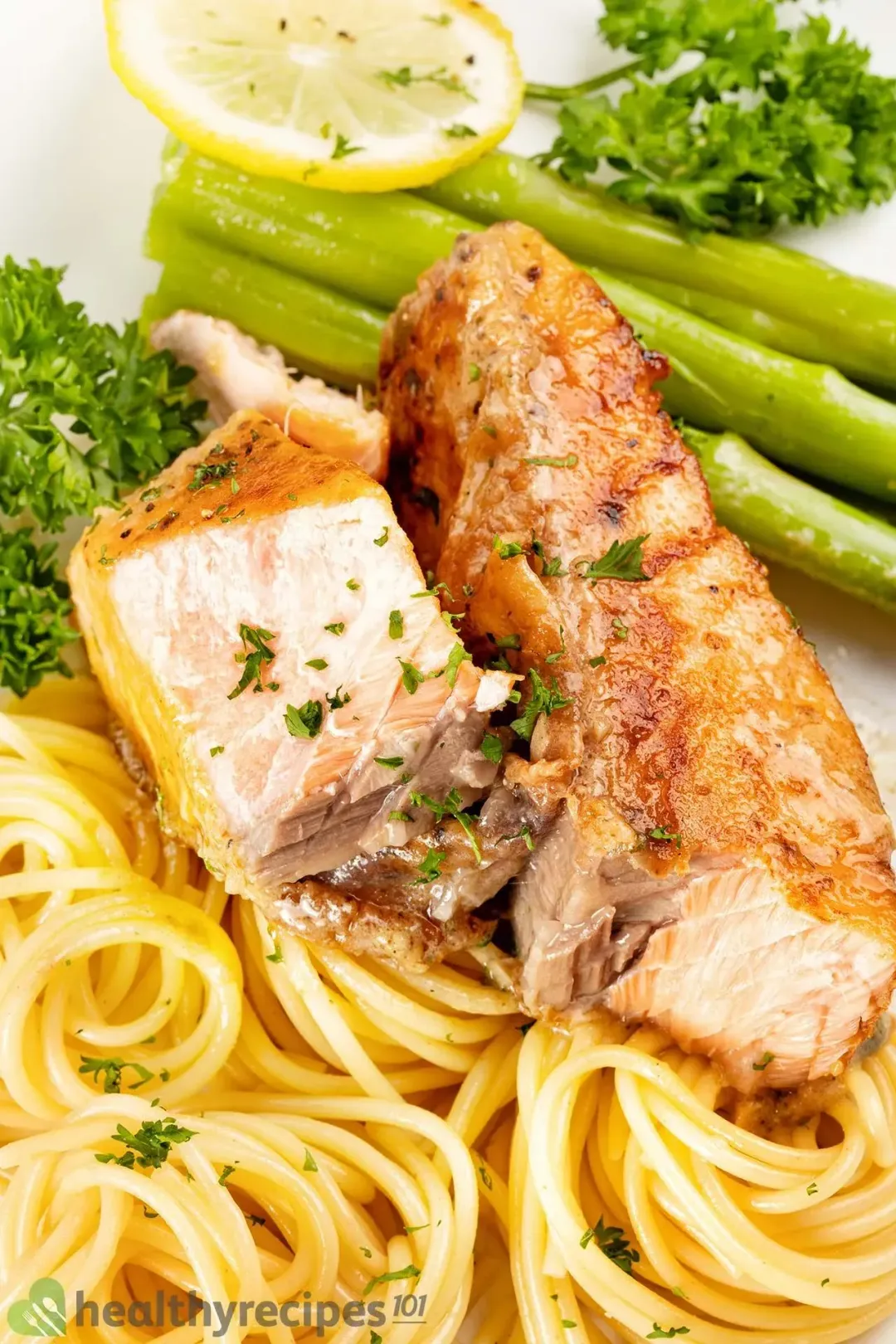 Two halves of a seared salmon filet sitting on a bed of cooked pasta and surrounded with cooked asparagus, a lemon slice, and parsley.