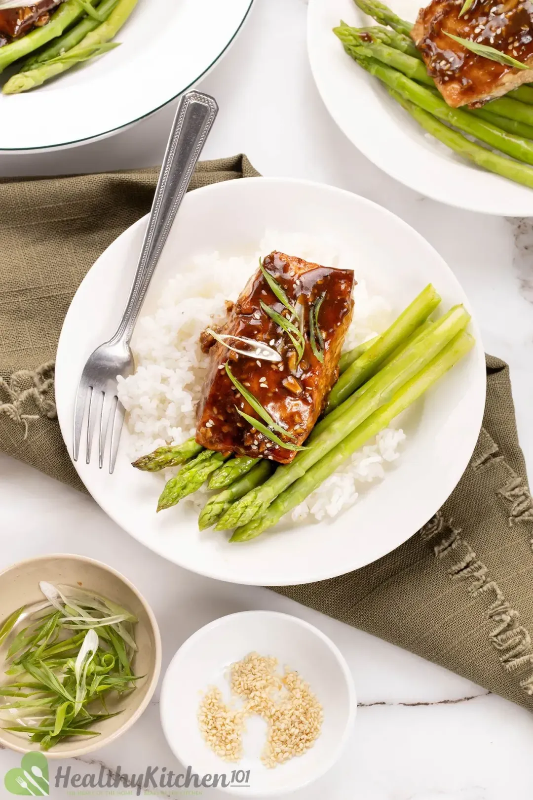 A serving plate of one baked teriyaki salmon filet served on cooked white rice and asparagus; on the side are two small bowls of roasted sesame seeds and chopped scallion.