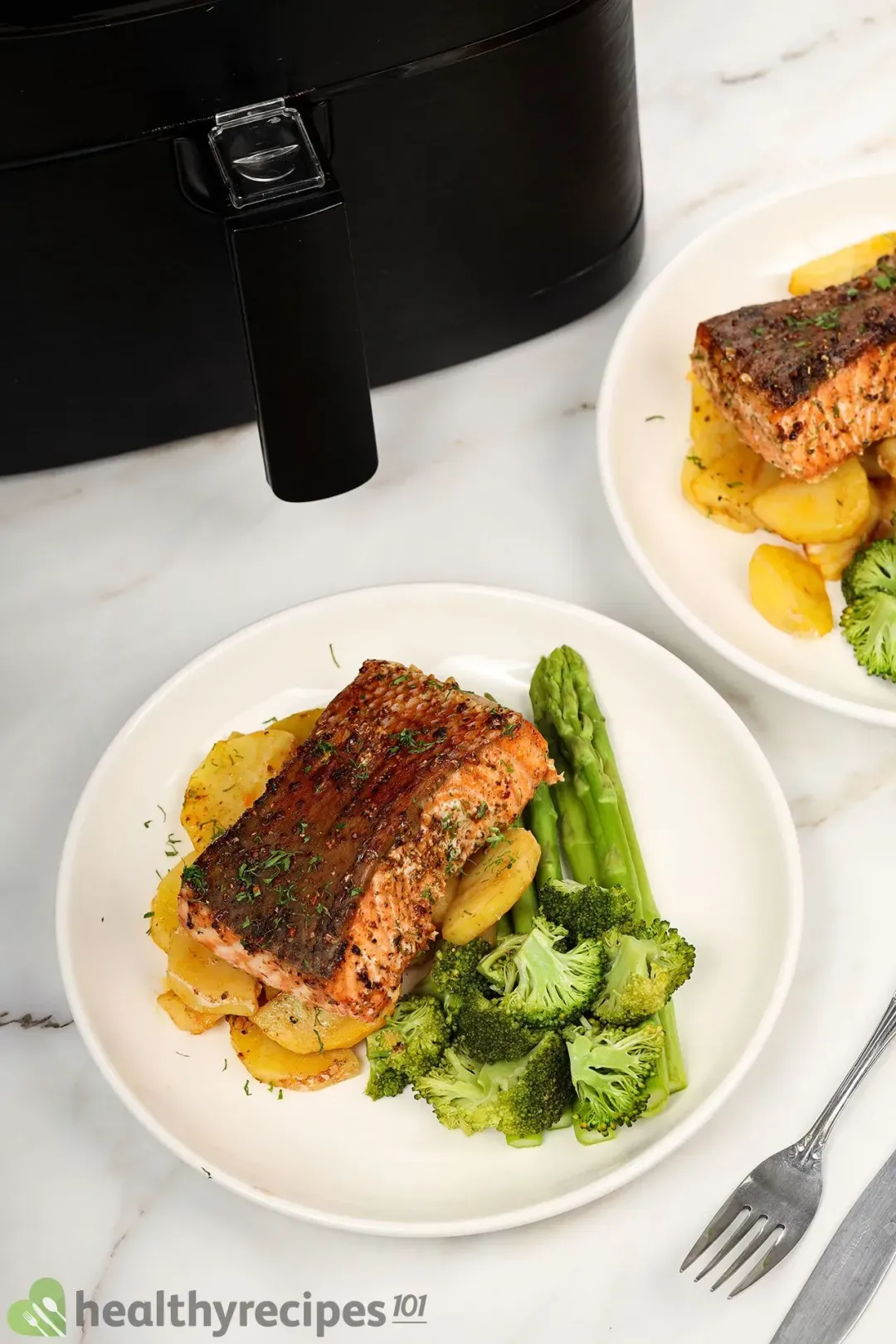 A round white plate containing potato slices, asparagus, broccoli florets, and a cooked salmon fillet laid near an air fryer