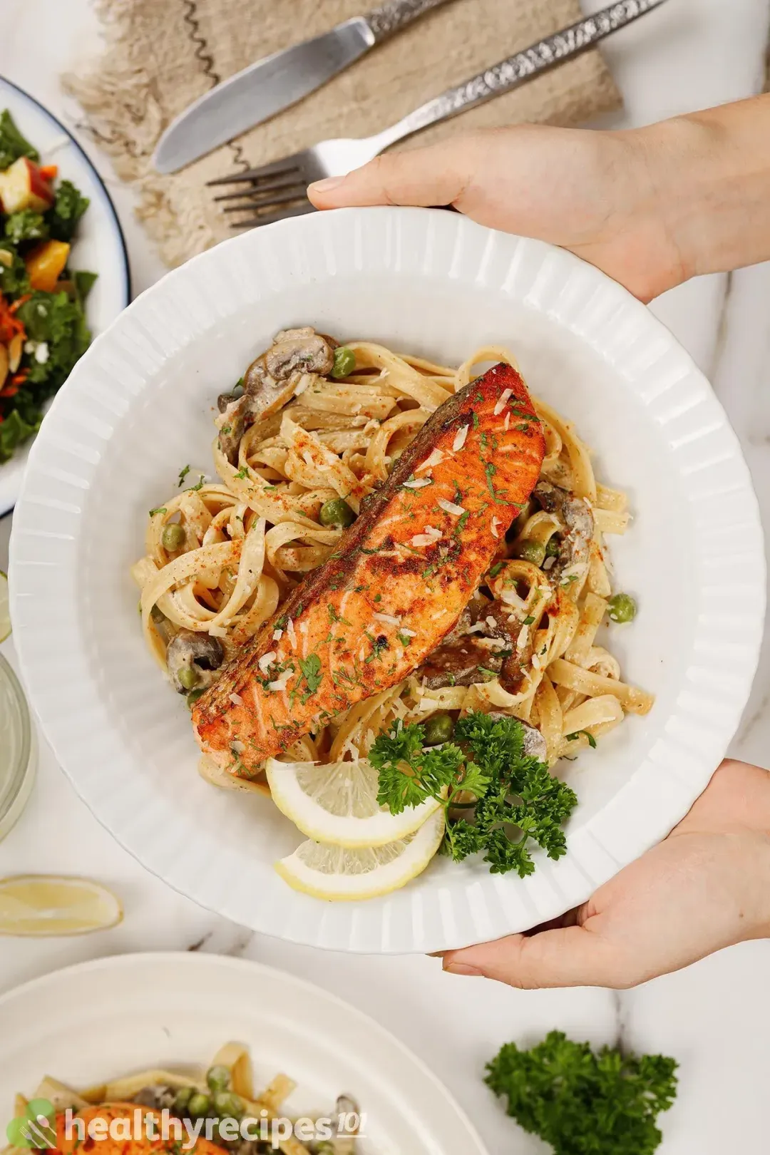 Two hands holding a plate of fettuccine pasta, a bright orange cooked salmon fillet, lemon slices, and fresh parsley