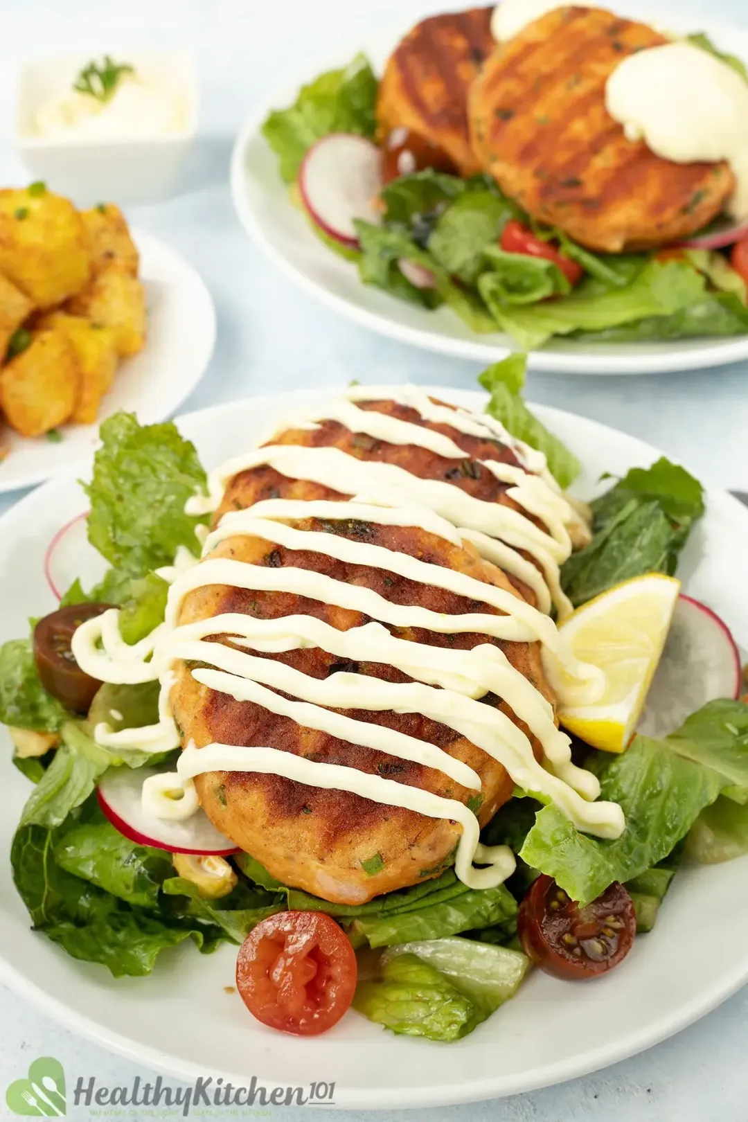 A picture of some salmon croquettes drizzled with the white sauce on a plate with fresh veggies and greens.