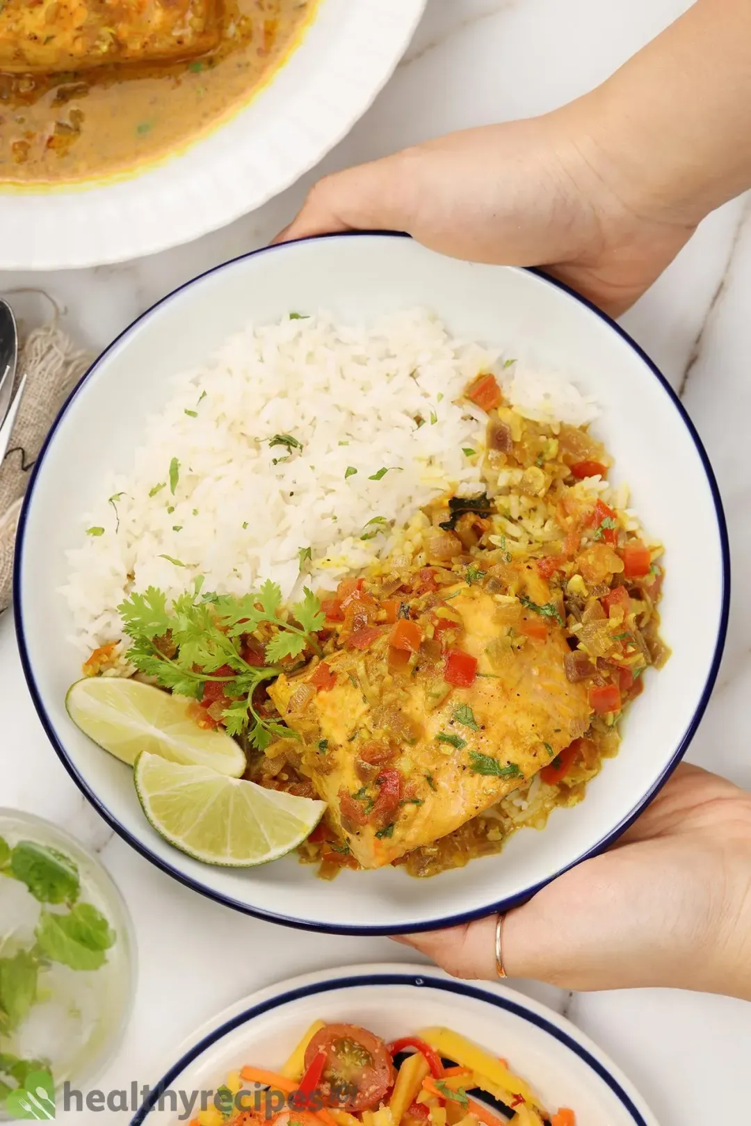 Two hands holding up a plate containing white rice, lime wedges, fresh coriander, a cooked salmon fillet covered in dark yellow curry and diced tomatoes
