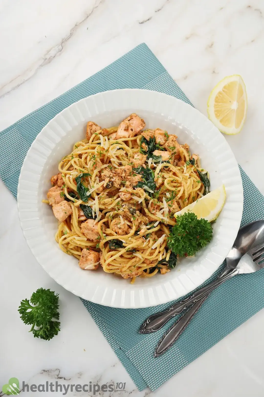A high-angle shot of a white deep plate of salmon pasta laid on a blue tablecloth and next to a lemon wedge, fresh, parsley, a fork, and a spoon