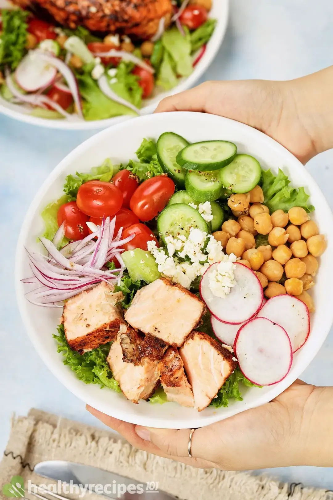 Two hands holding a plate of salad consisting of lettuce, chickpeas, sliced radish, sliced cucumbers, cherry tomatoes, red onion, and pan-seared salmon cubes
