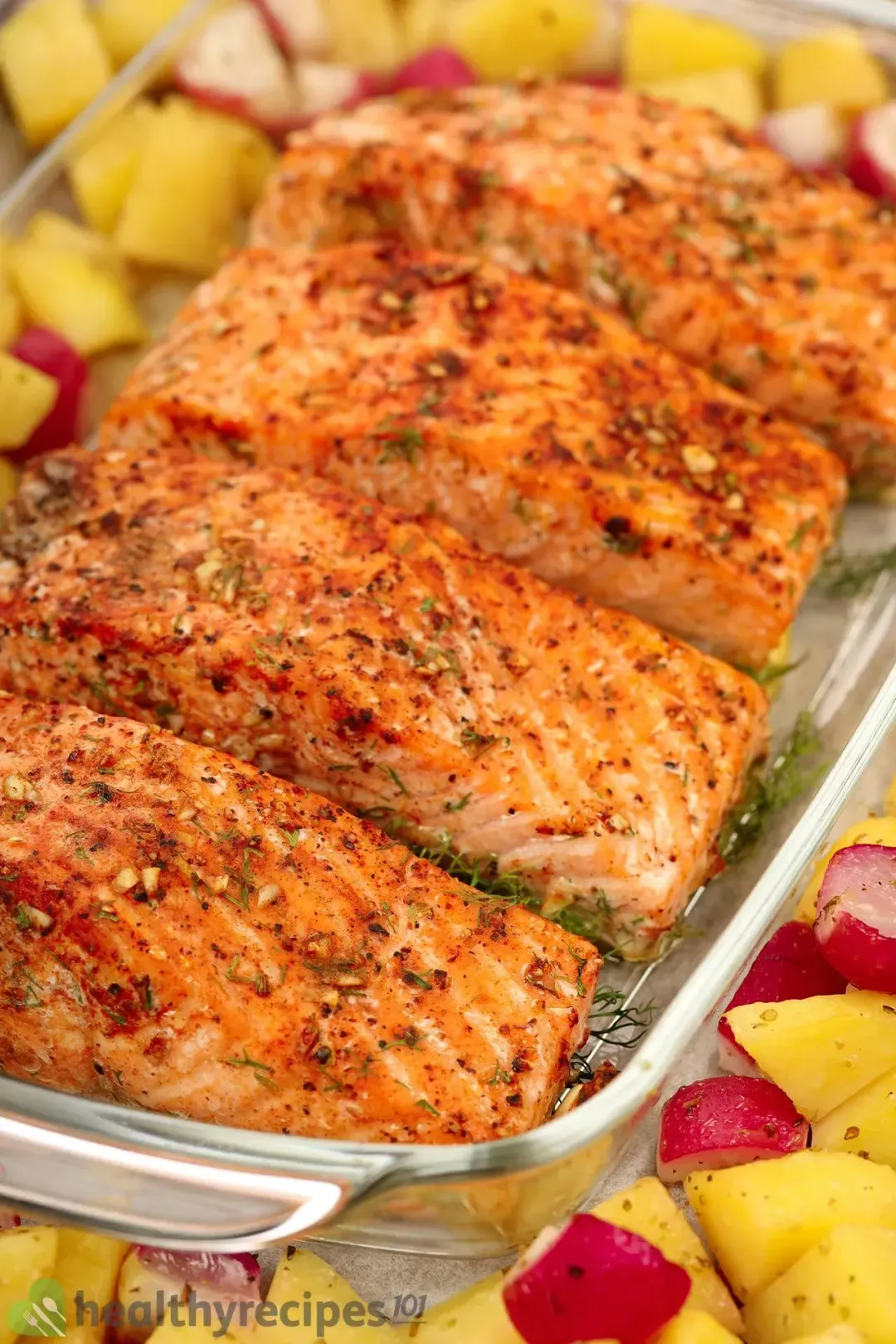 A large glass tray containing four baked salmon fillets surrounded by potato cubes and red radish cubes