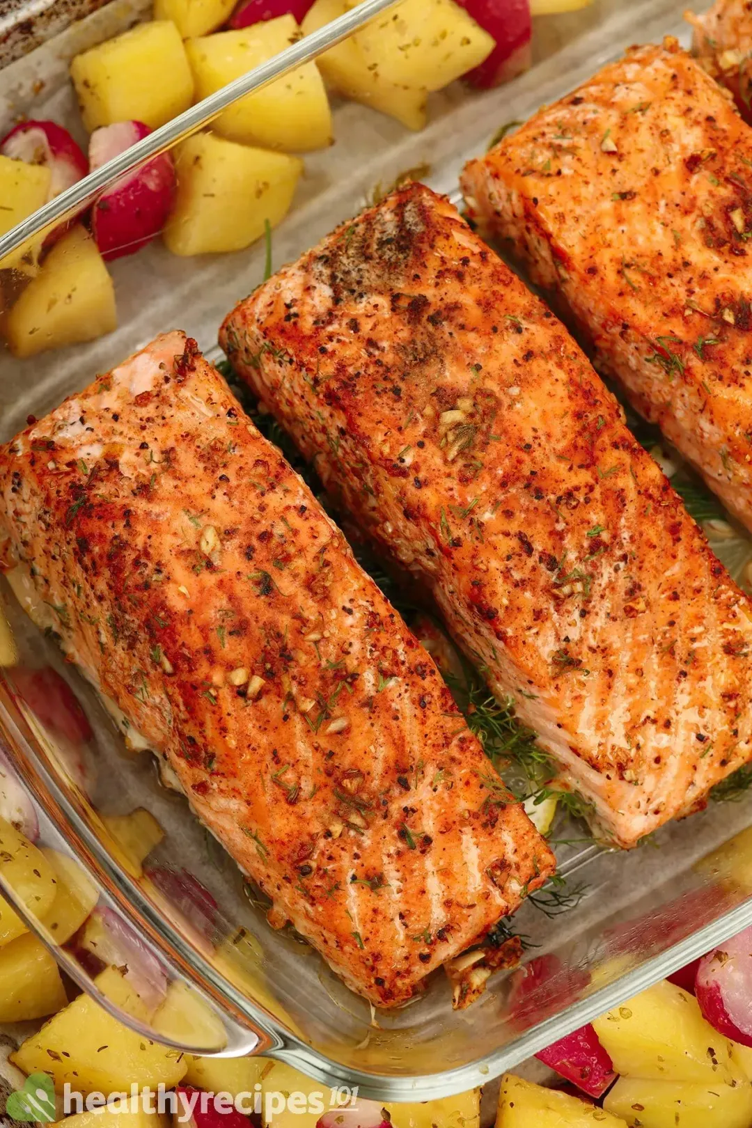 A close-up shot of baked salmon fillets laid in a glass tray surrounded by potato cubes and red radish cubes