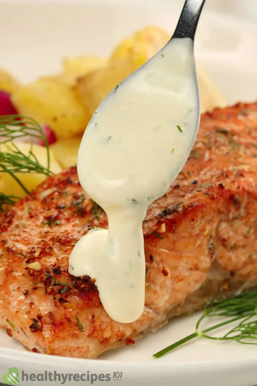 A close-up shot of a spoon slowly dropping a thick and creamy sauce onto a cooked salmon fillet with potatoes and dill in the background