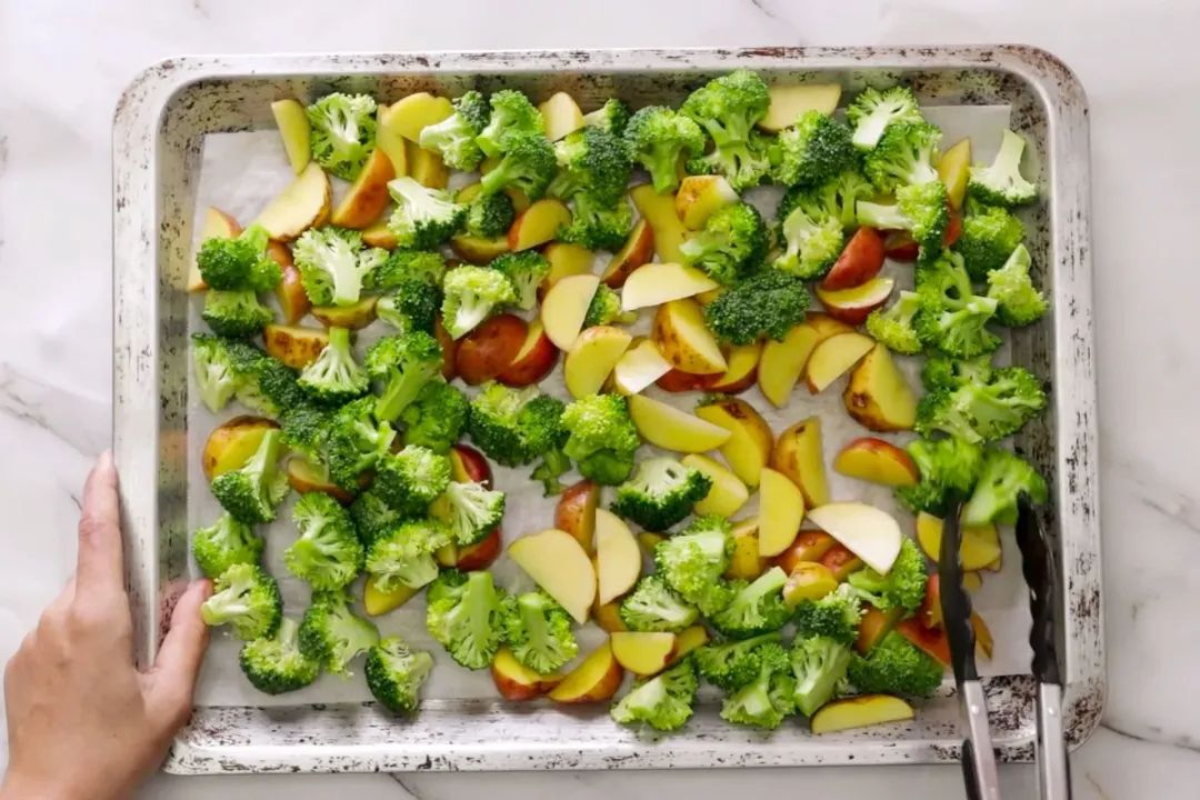 A baking sheet lined with parchment paper, on which there are raw potato wedges and raw broccoli pieces being arranged in a single layer.