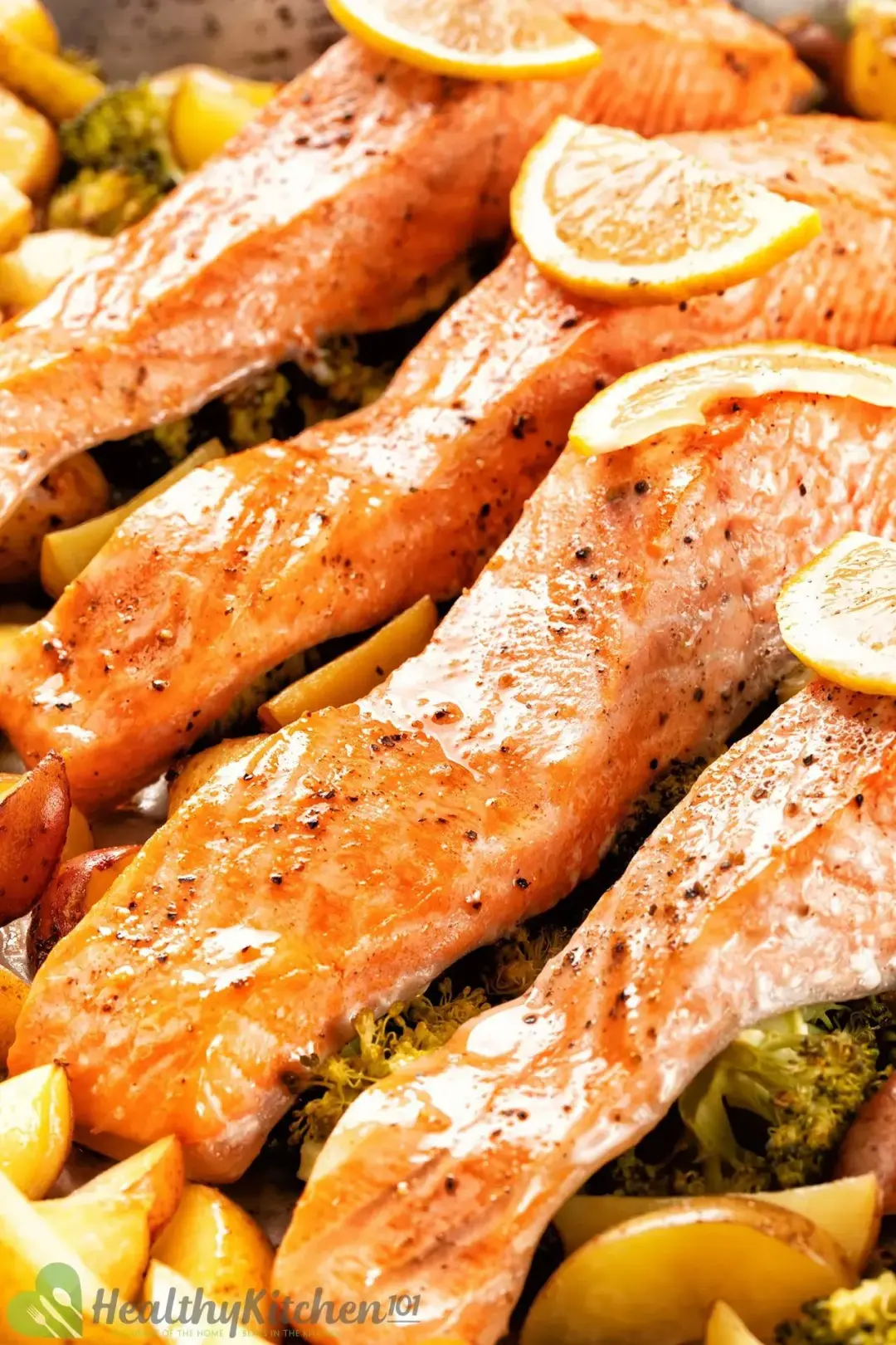 A close-up shot of our baked salmon filets are topped with lemon slices and assembled on a baking sheet with baked potatoes and broccoli.