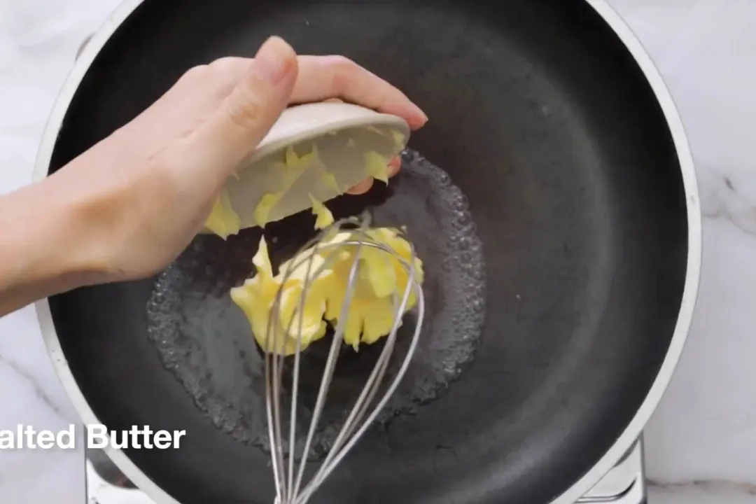 A scoop of butter is being removed from a small bowl held by a hand using an egg whisker and added to a skillet.