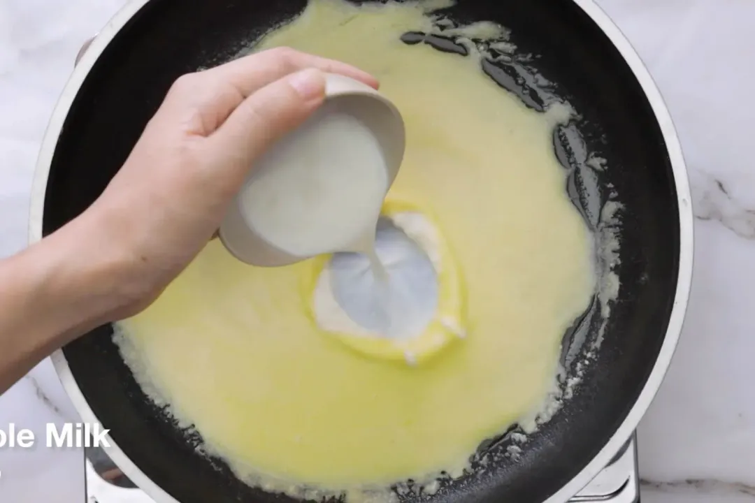 A hand pouring a milky mixture from a small bowl to a skillet filled with melted butter.