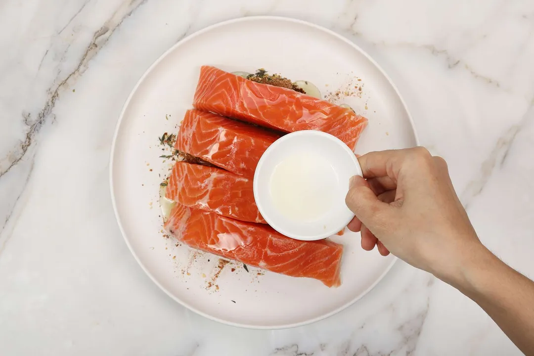 a hand holds a small bowl of oil on top of a salmon fillet plate