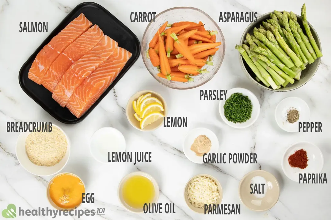 A black dish containing four raw salmon fillets, a bowl of baby carrots, a bowl of asparagus stalks, and a bowl of lemon slices laid near various condiments