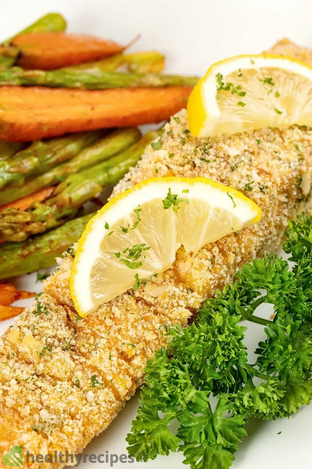 A close-up shot of a breadcrumb-crusted salmon fillet surrounded by lemon pieces and parsley with carrots and asparagus in the blurred background