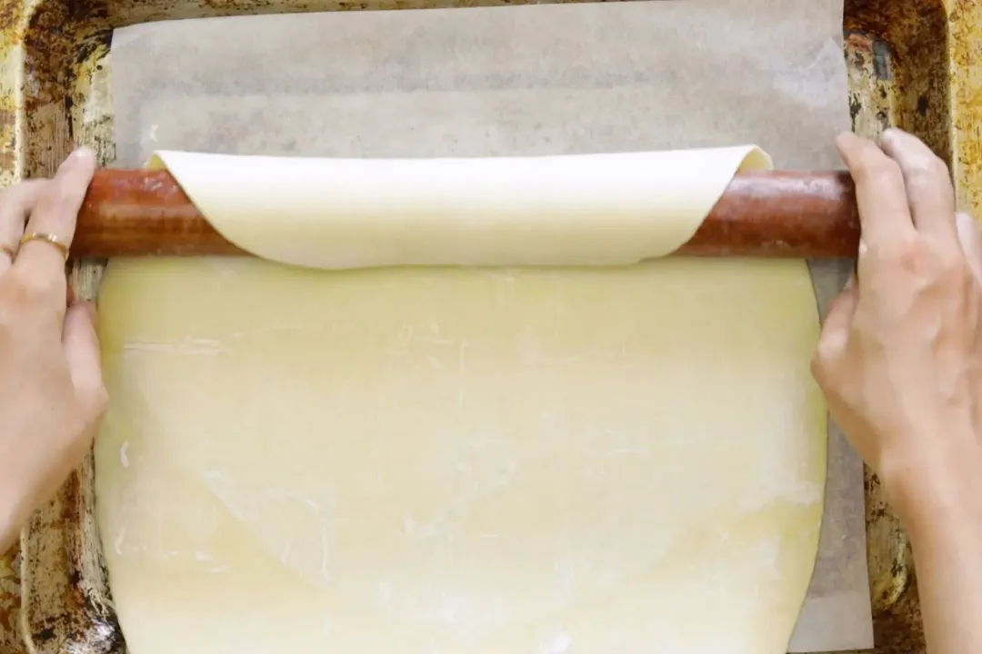 Two hands rolling a puff pastry dough on a baking pan lined with parchment paper