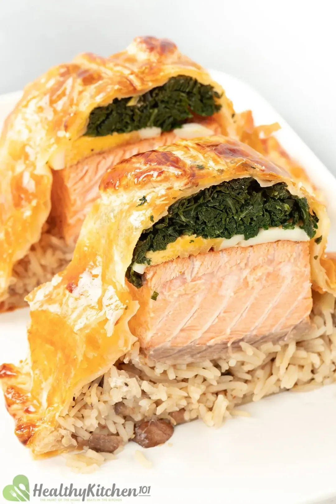 Two slices of salmon coulibiac with layers of cooked kale, boiled eggs, salmon, and mushroom rice on a white plate