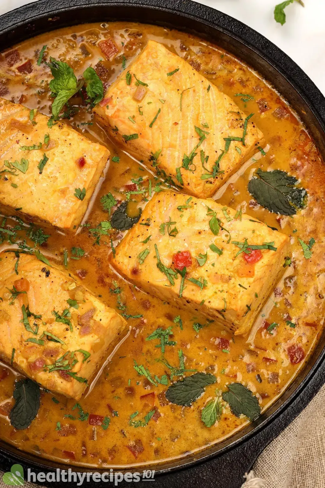 A skillet containing dark yellow curry with four rectangle salmon fillets and small pieces of red onions