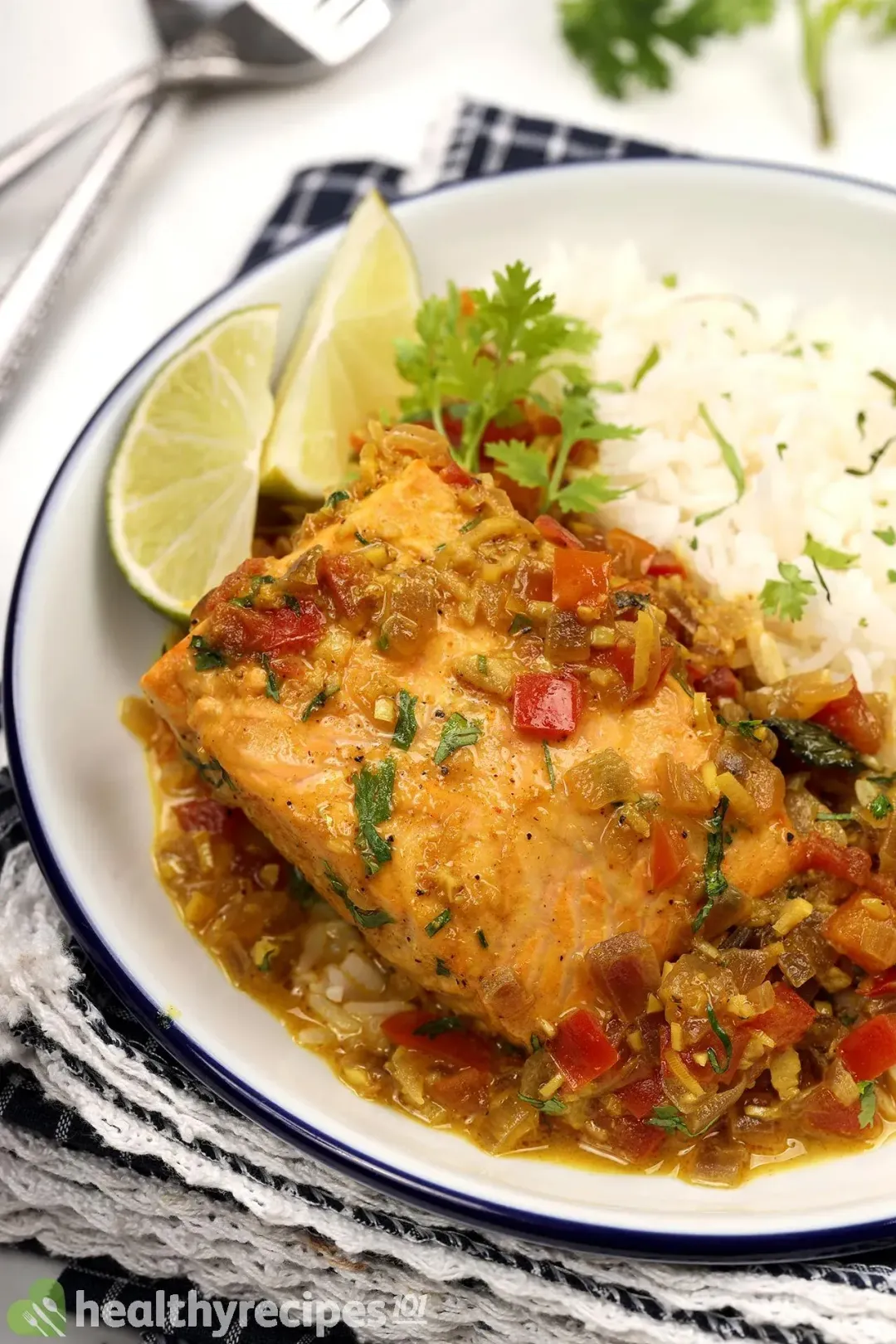 A close-up shot of white rice, lime wedges, and a cooked salmon fillet covered in yellow curry, chopped herbs, and diced tomatoes