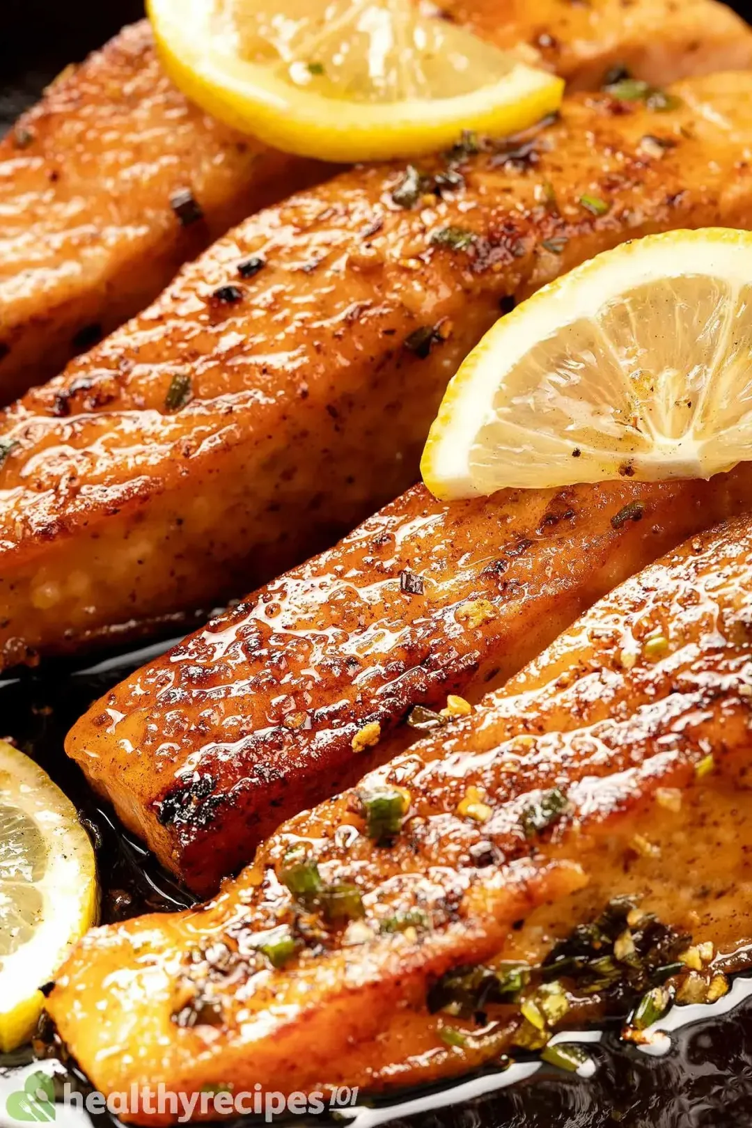Four salmon filets coated in a glossy butter sauce, garnished with lemon slices in a cast iron skillet