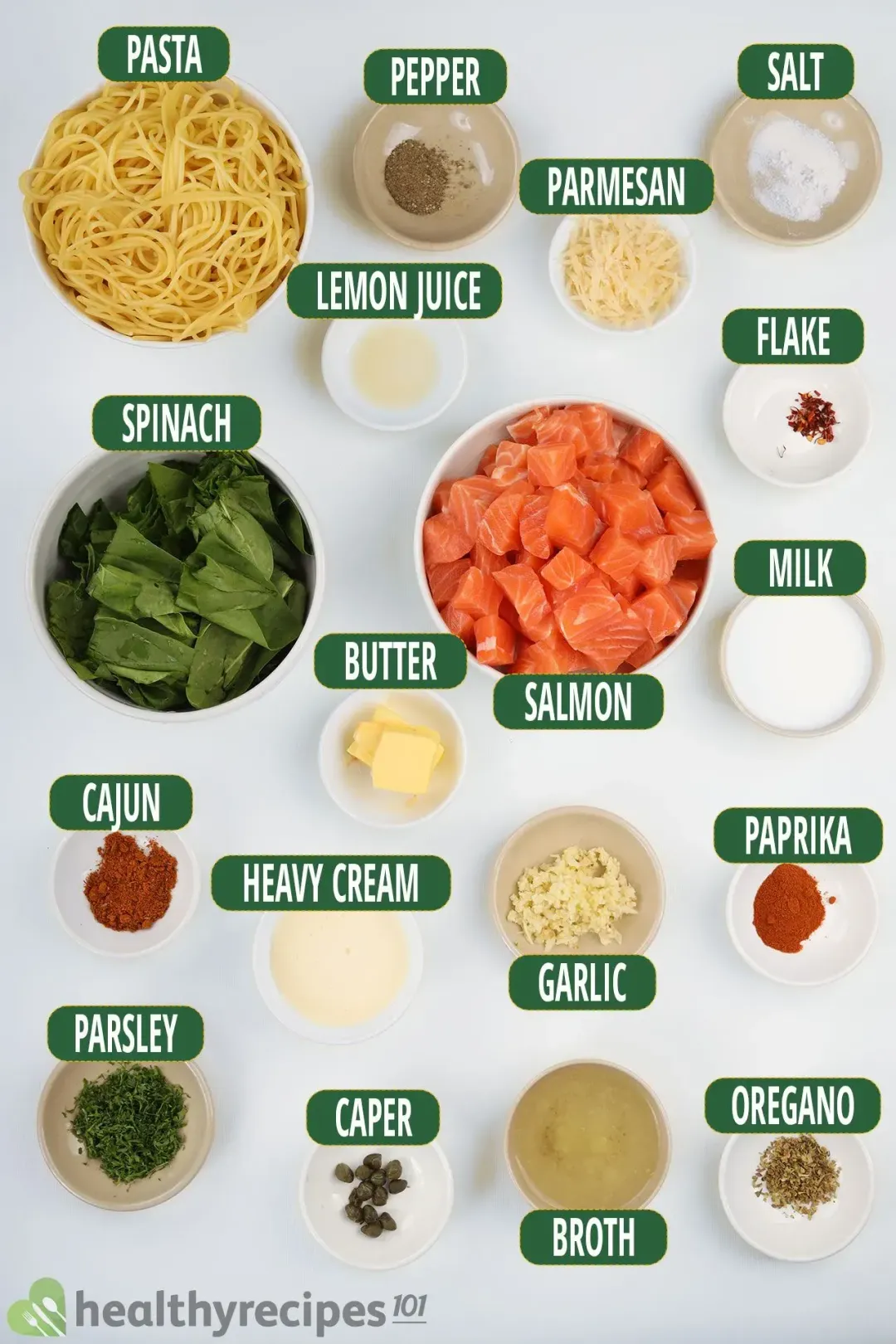 A bowl of raw cubed salmon fillets, a bowl of spinach, and a bowl of cooked spaghetti laid near various condiments