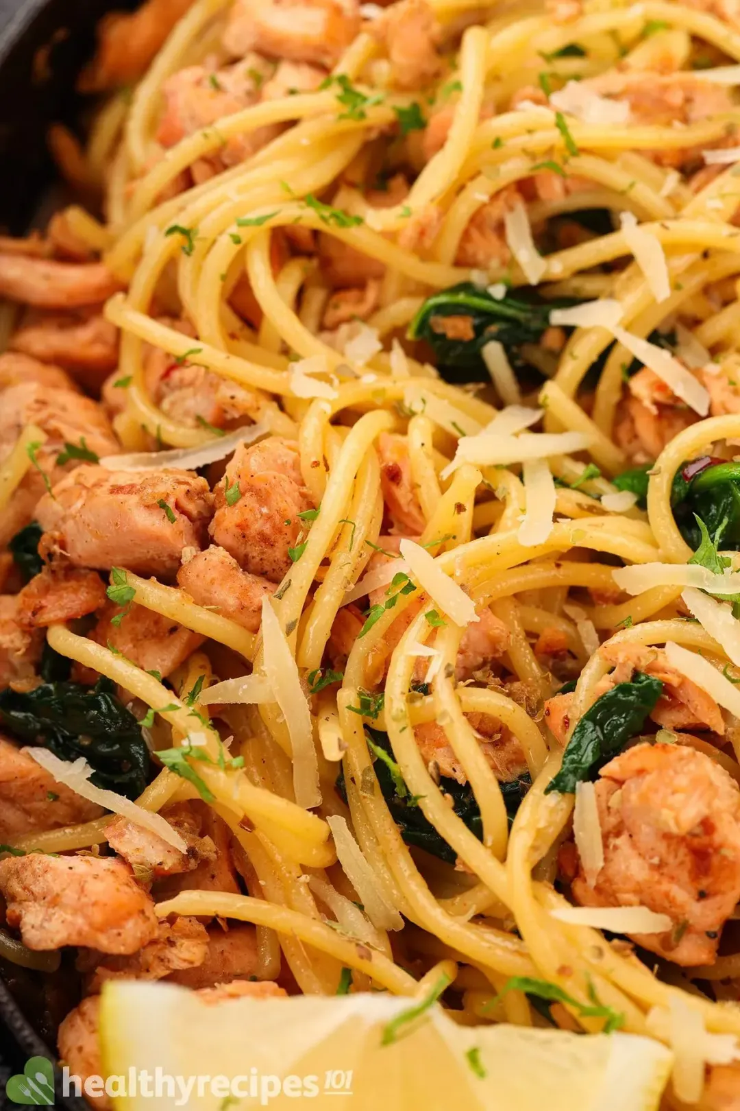 A close-up shot of a salmon pasta featuring spaghetti, cooked salmon cubes, shredded parmesan cheese, and cooked spinach