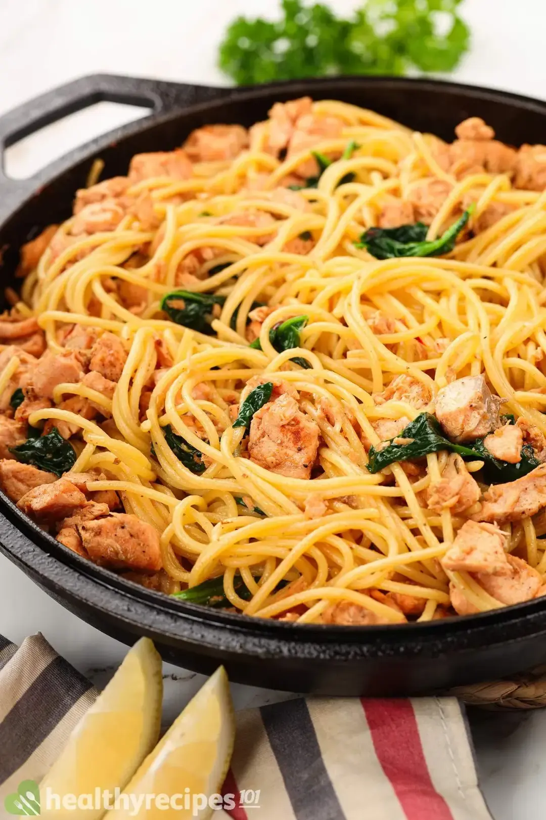 A skillet filled with spaghetti, cooked salmon cubes, and cooked spinach laid near a striped tablecloth, two lemon wedges, and fresh parsley
