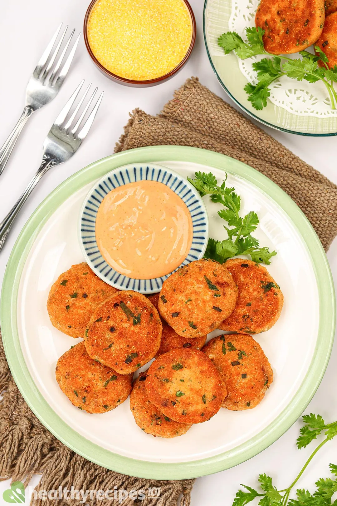 a plate of salmon patties with a small bowl of yellow sauce garnish with fork, a bowl of cornmeal and coriander