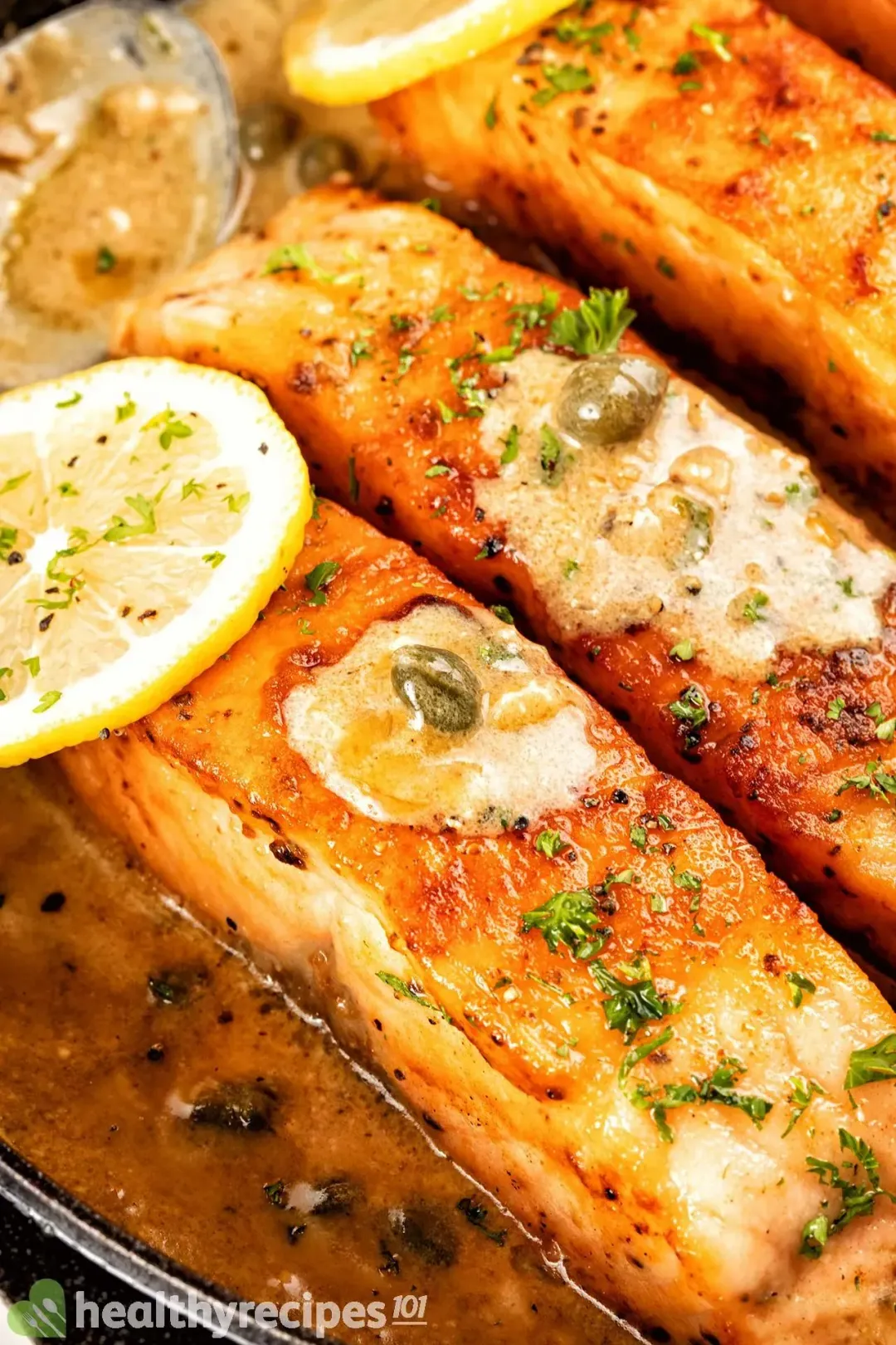 A close-up view of goldenly seared salmon filets sitting in a cooking juice, on top of which there are capers, lemon slices, and a creamy sauce.
