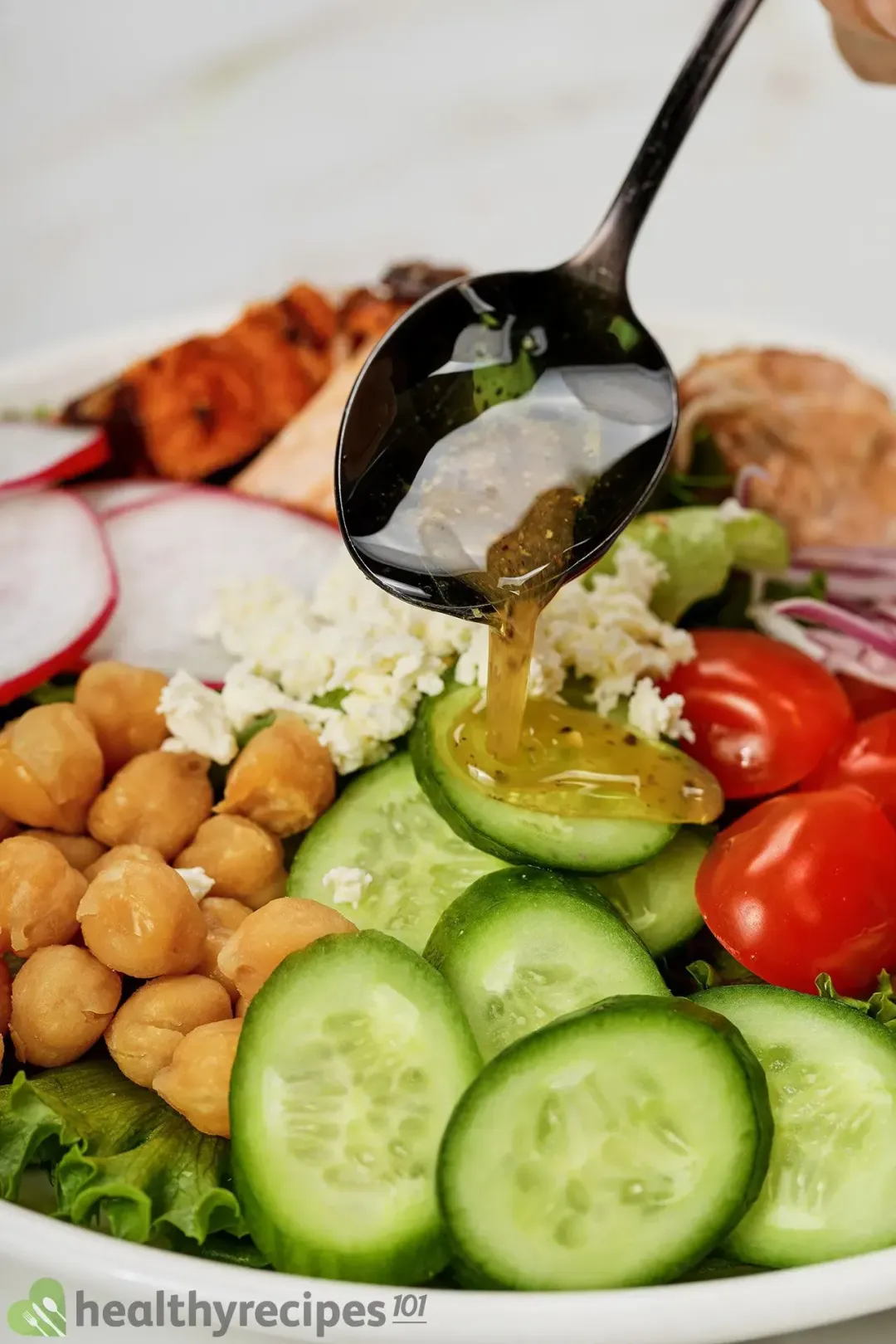 A black spoon dispensing a thick and syrupy dressing onto slices of cucumbers, chickpeas, and cherry tomatoes of a salad