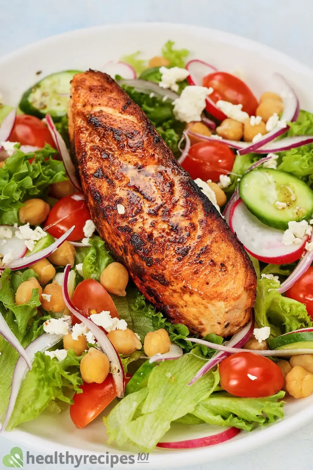 A pan-seared salmon fillet with charred marks laid over a bed of lettuce, chickpeas, halved cherry tomatoes, sliced radish and sliced cucumbers