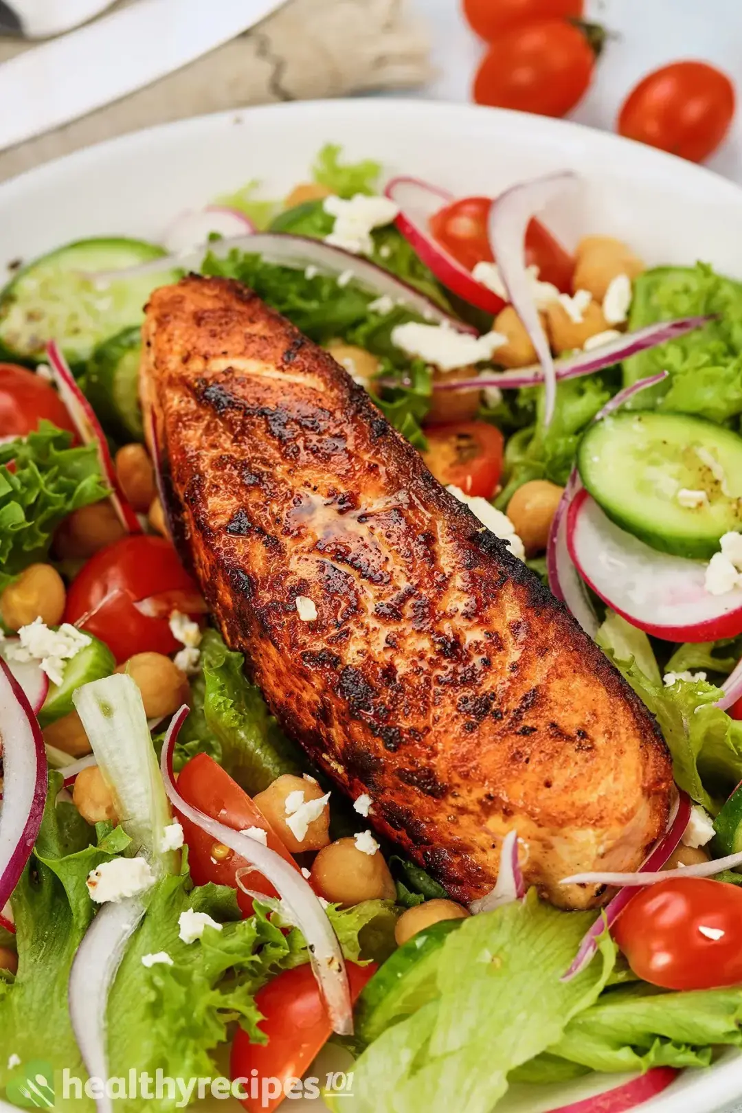 A pan-seared salmon fillet with charred marks laid over a bed of lettuce, chickpeas, halved cherry tomatoes, sliced radish and sliced cucumbers