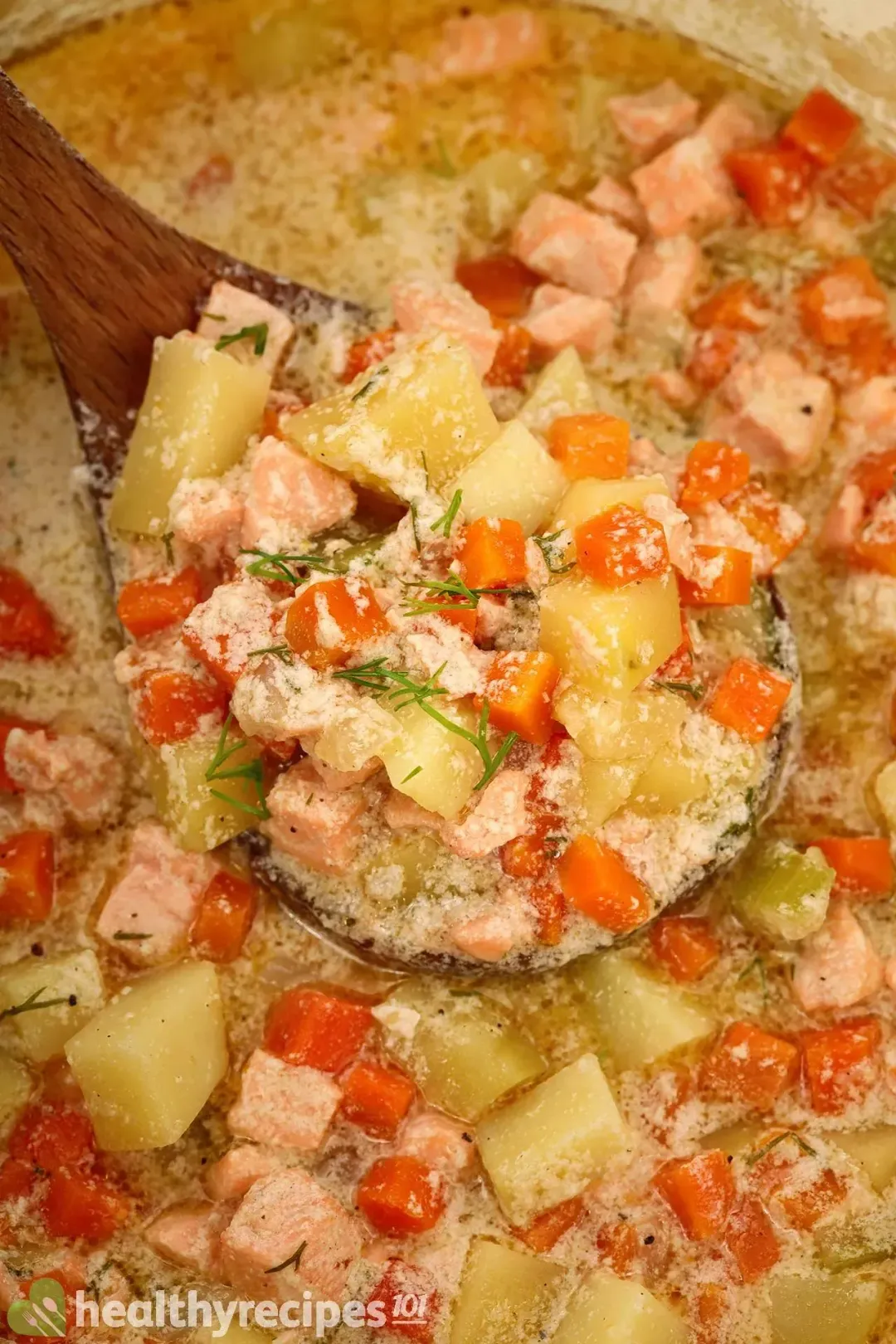 A ladle holding up coarsely-diced potatoes, coarsely-diced carrots, and salmon cubes in a pot of soup