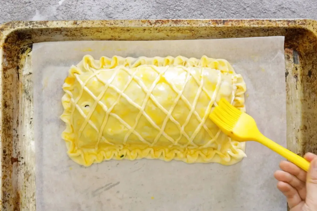 A hand using a butter brush to spread butter all over the surface of a large and uncooked puff pastry