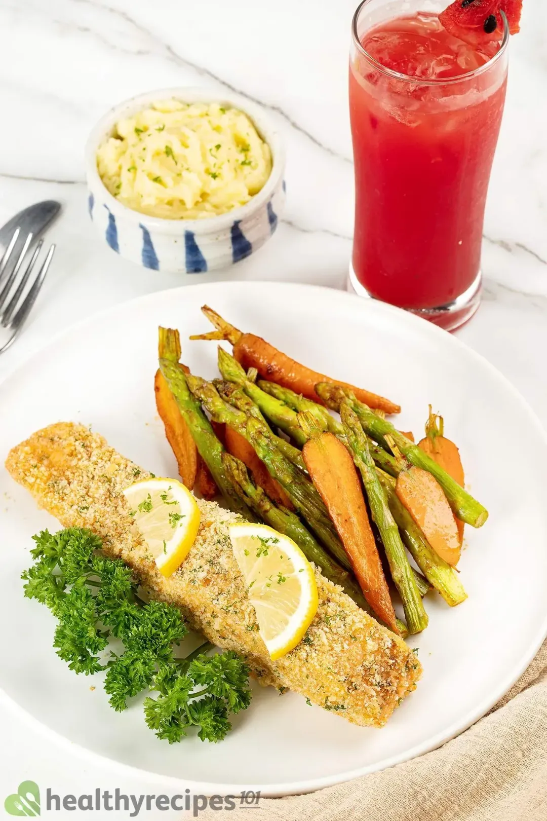 A white plate containing a salmon fillet crusted with breadcrumbs, some baby carrots, asparagus stalks, two lemon pieces, and parsley laid next to mashed potatoes and watermelon juice