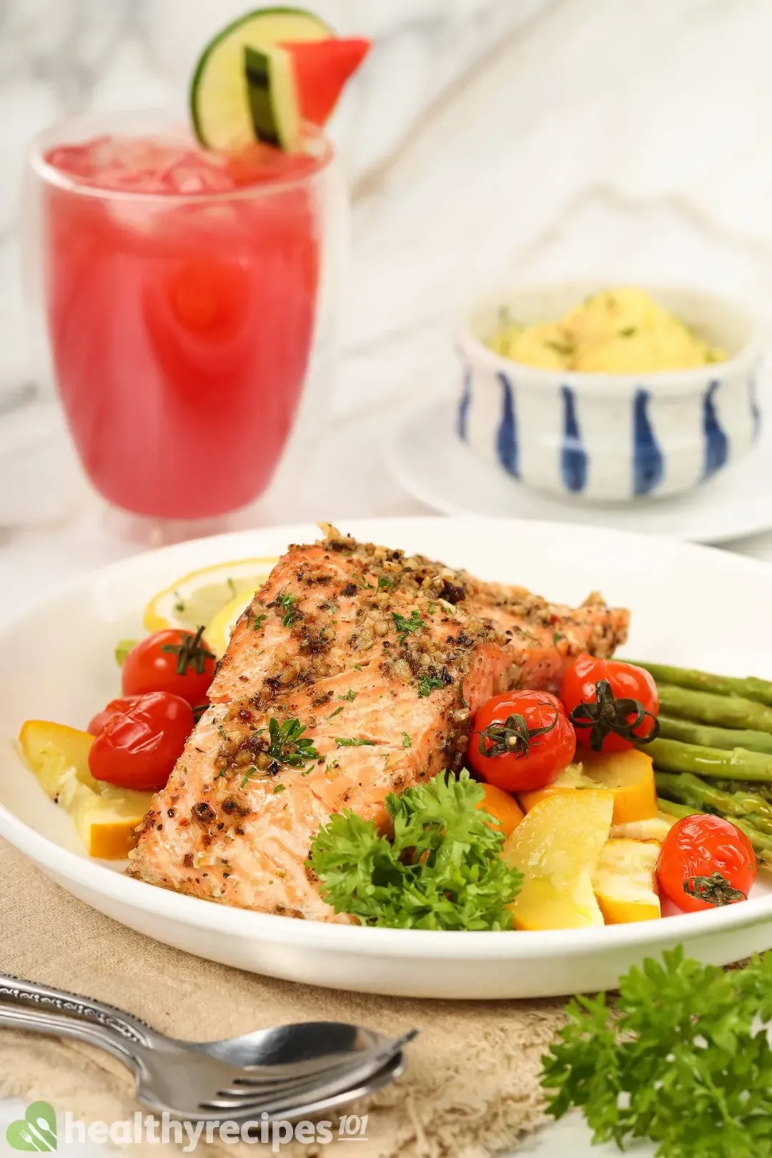 A plate of baked salmon placed on a small tablecloth with a glass of lime watermelon juice and a bowl of mashed potatoes on the background