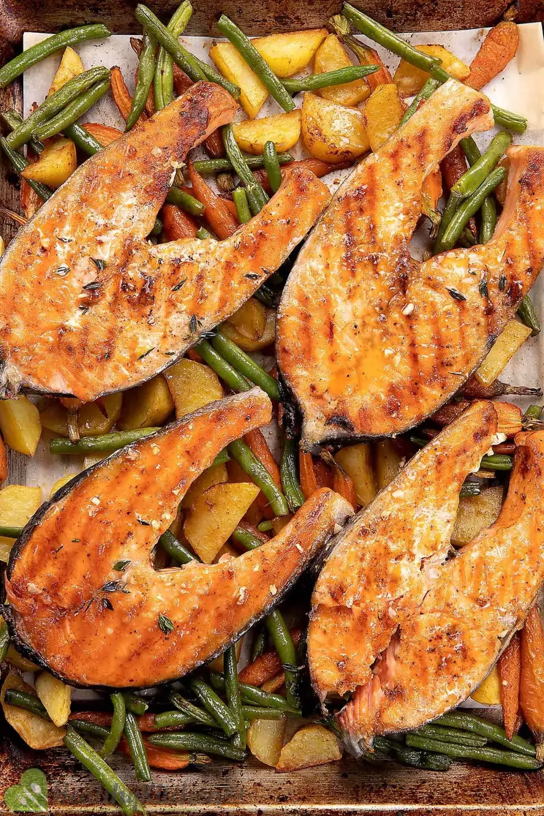 Four salmon steaks with charred marks sitting atop a tray of roasted baby potatoes, carrots, and string beans