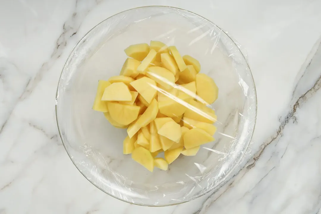 A large glass bowl containing uncooked potato cubes covered with a cling wrap