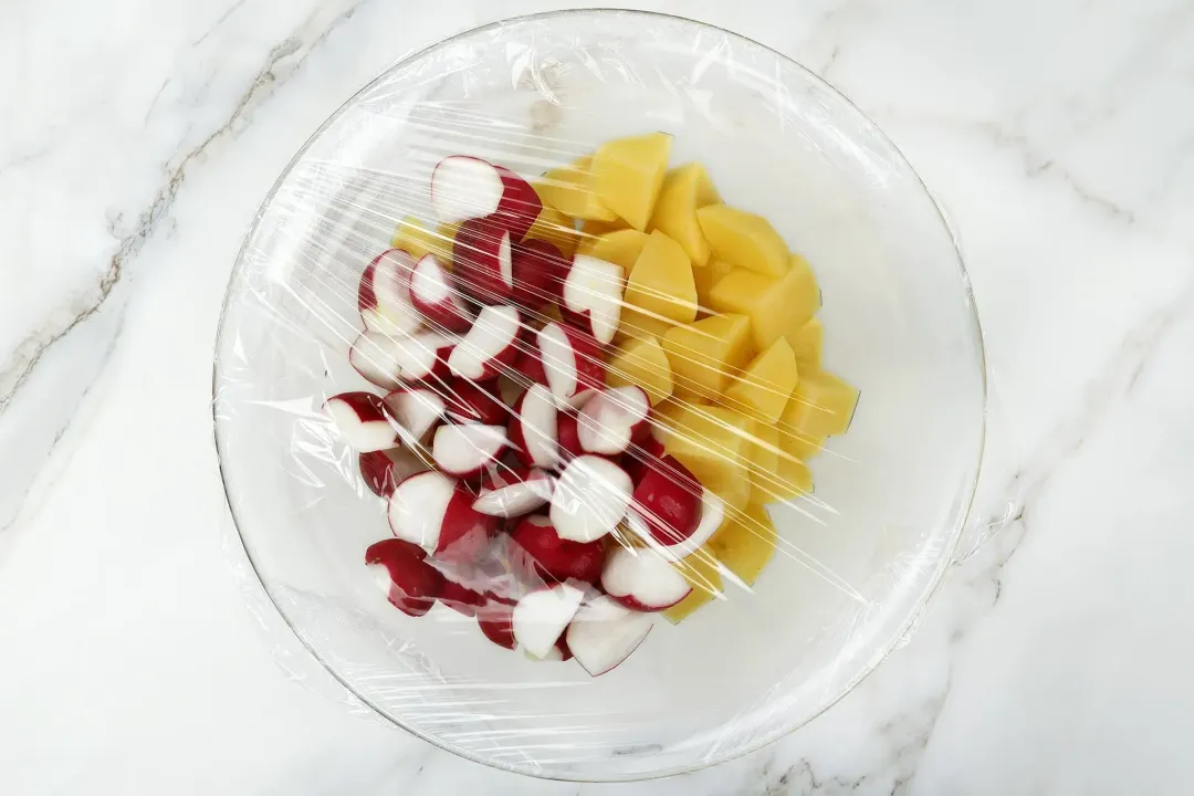 A large glass bowl containing potato cubes and red radish cubes covered in cling wrap