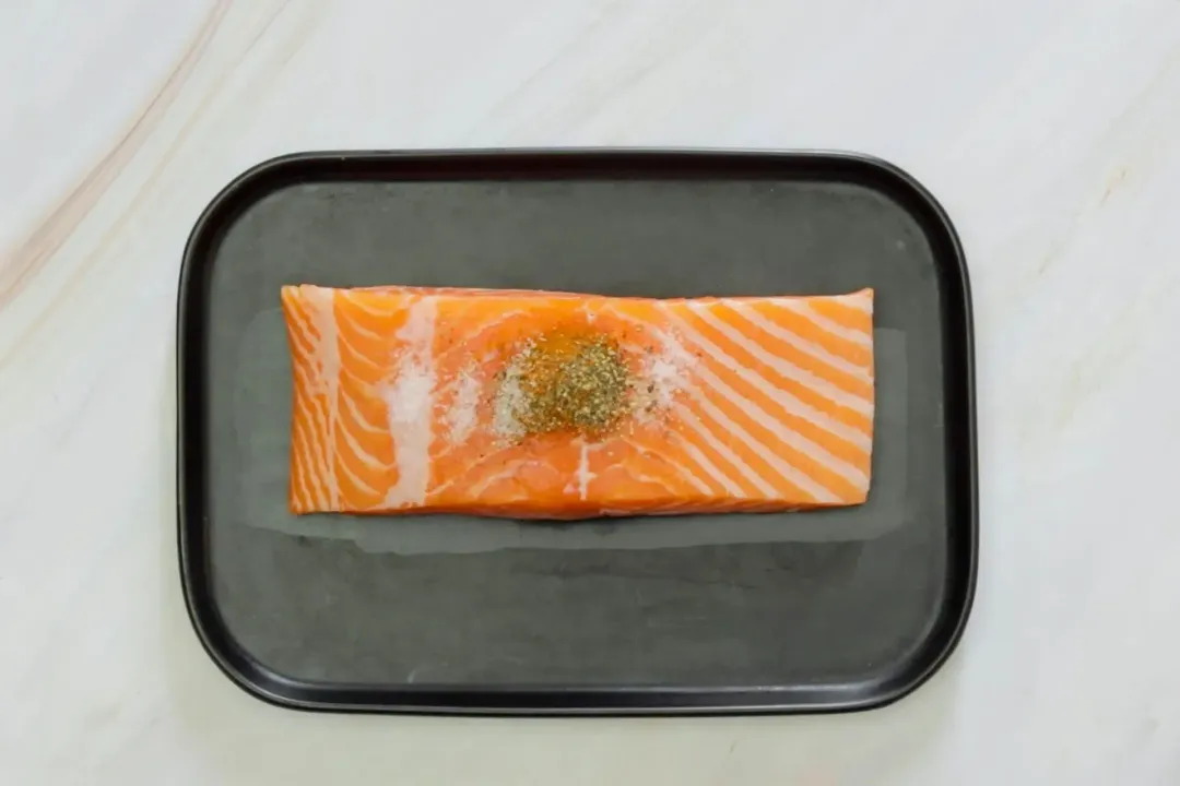 A black rectangle plate containing a large salmon fillet with some spices laid on top