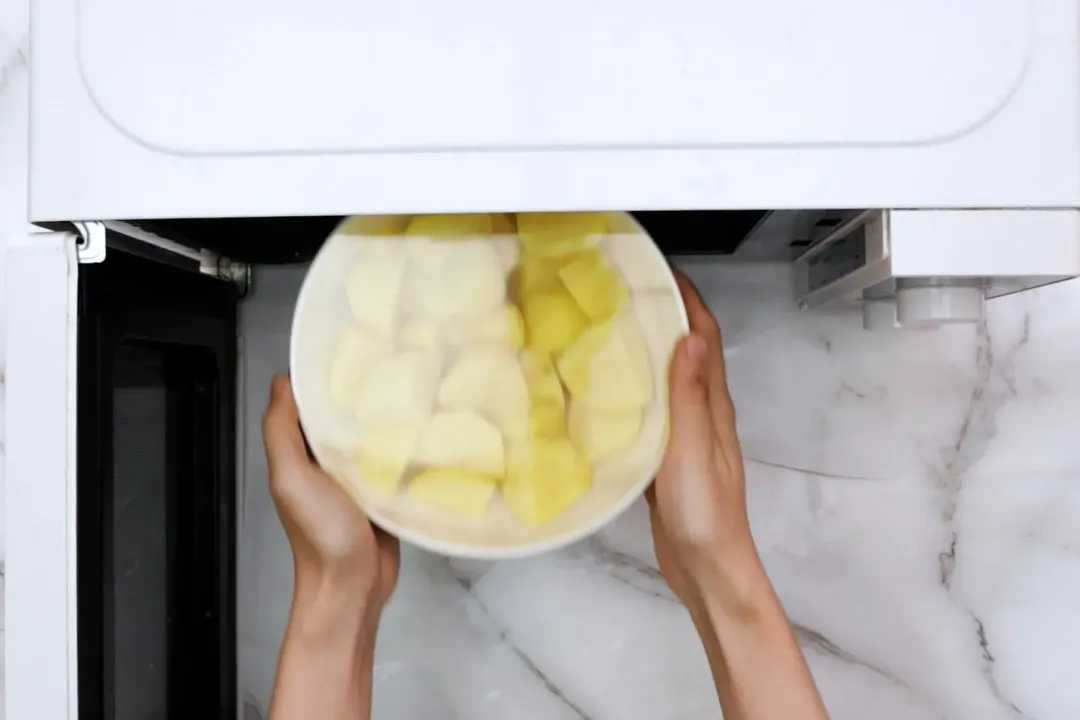 Two hands putting a bowl of potato wedge covered in plastic wrap into a microwave