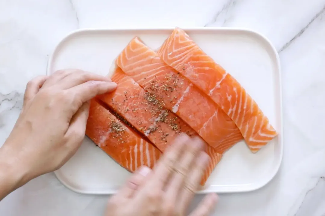 Four raw salmon filets are being rubbed with a spice mix by hand.