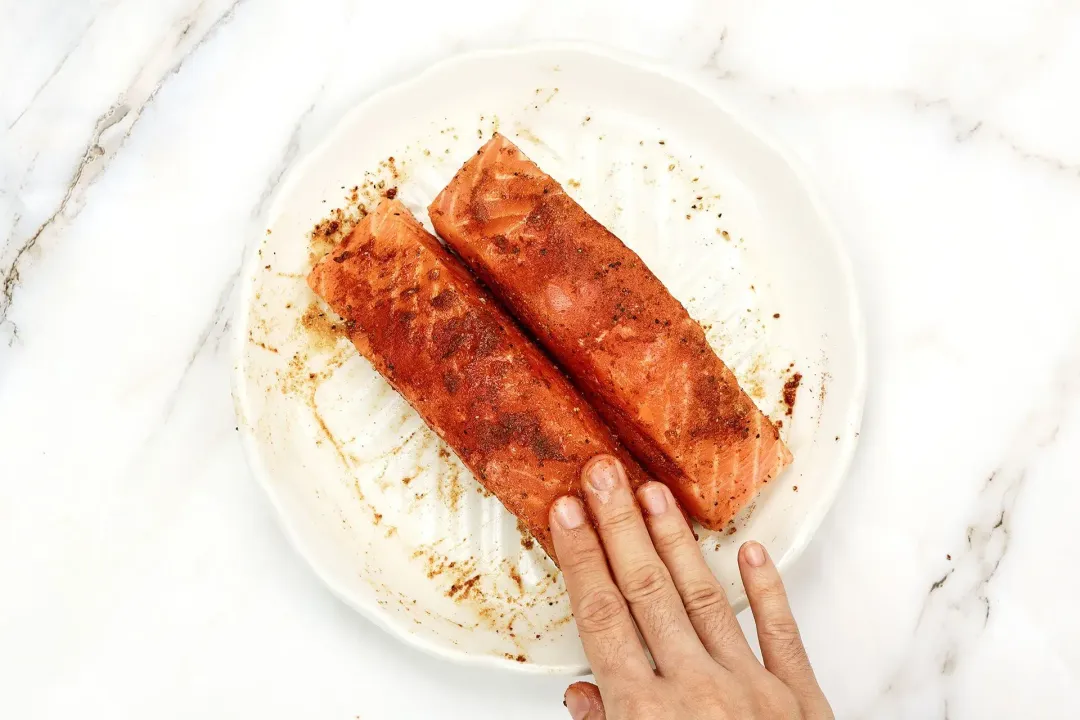 A hand dry-rubbing spices onto two raw salmon fillets laid on round white plate