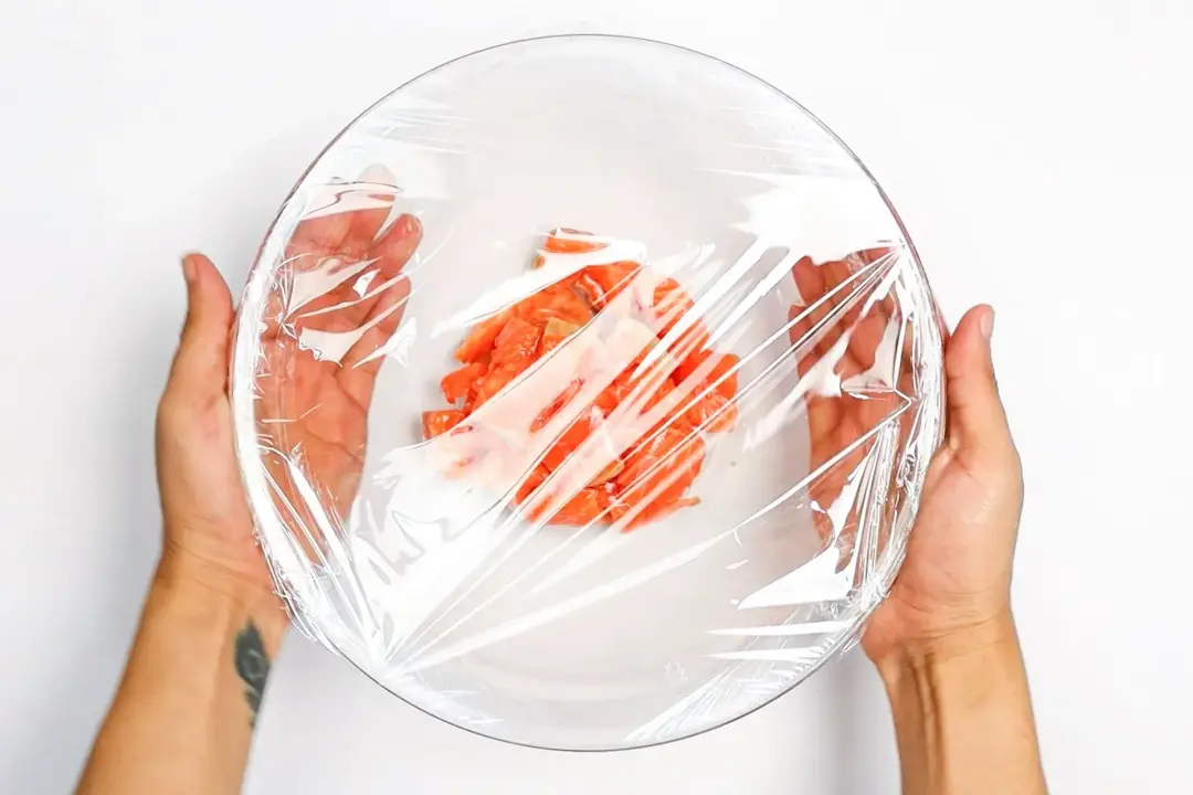 A picture of two hands holding a glass bowl covered in plastic wrap with some orange salmon cubes inside.