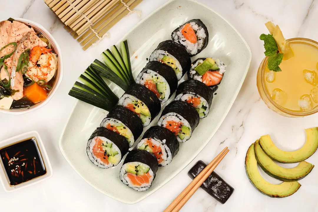 a plate of sushi decorated with a glass of juice and a bowl of soup with avocado slices