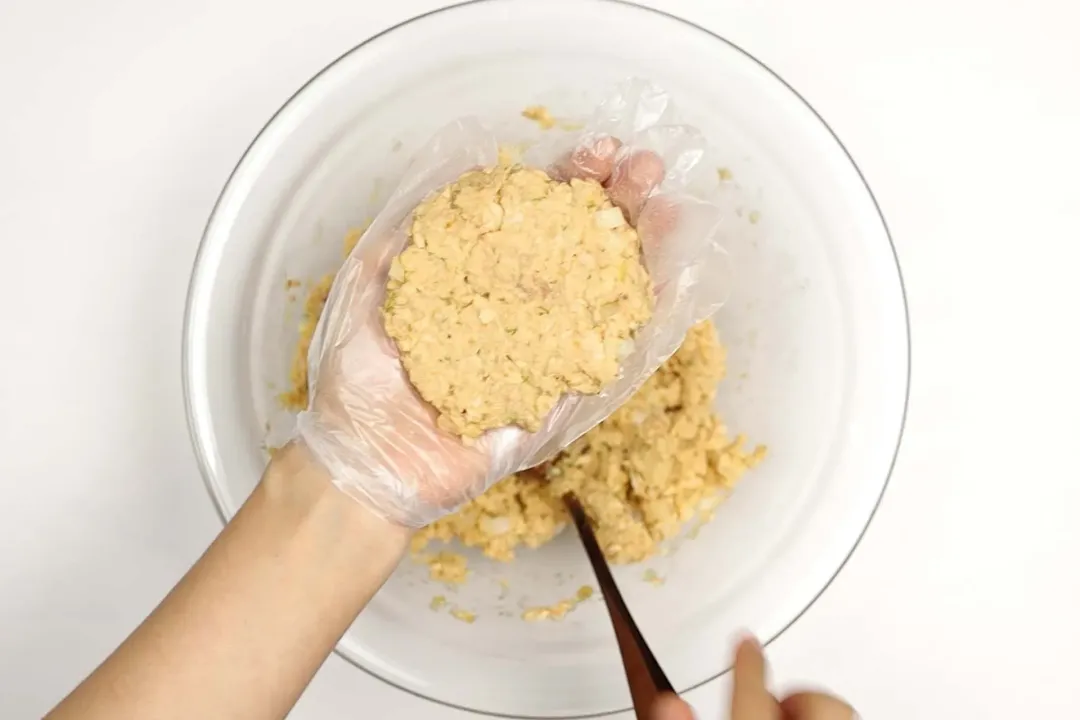 A gloved hand holding a light brown pattie mixture over a glass bowl containing the same mixture